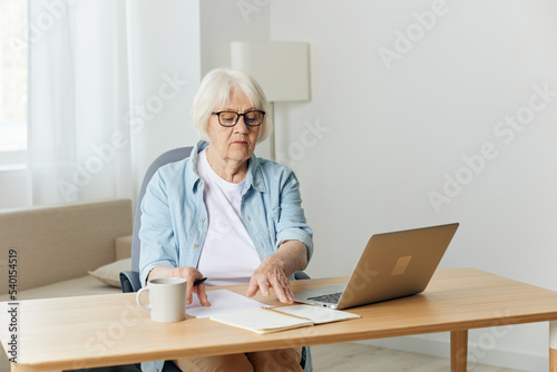 a serious, focused, elderly woman sits at home in a stylish shirt and analyzes the work making notes in her notebook, taking the matter seriously. Working from home for the elderly