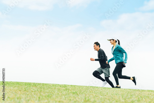青空の見える公園でランニングするスポーツウェアを着たアジア人の男女（走る） 