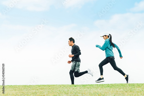青空の見える公園でランニングするスポーツウェアを着たアジア人の男女（走る） 