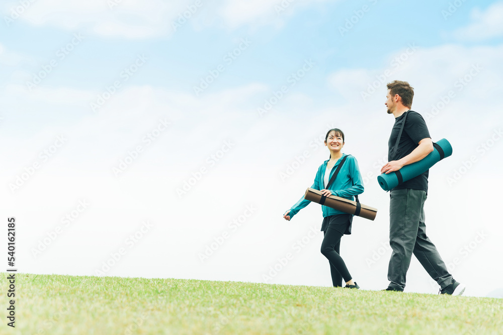 青空の見える公園をヨガマットを持って歩くスポーツウェアの男女（ヨガ教室）
