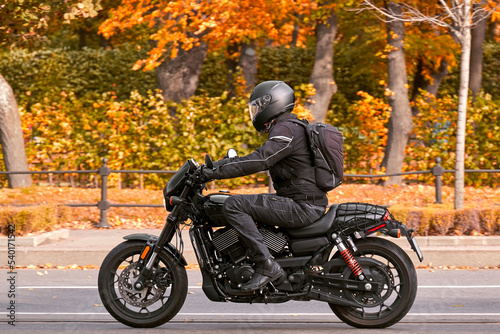 A motorcyclist rides a black retro motorcycle on the road next to the autumn park, the end of the season.