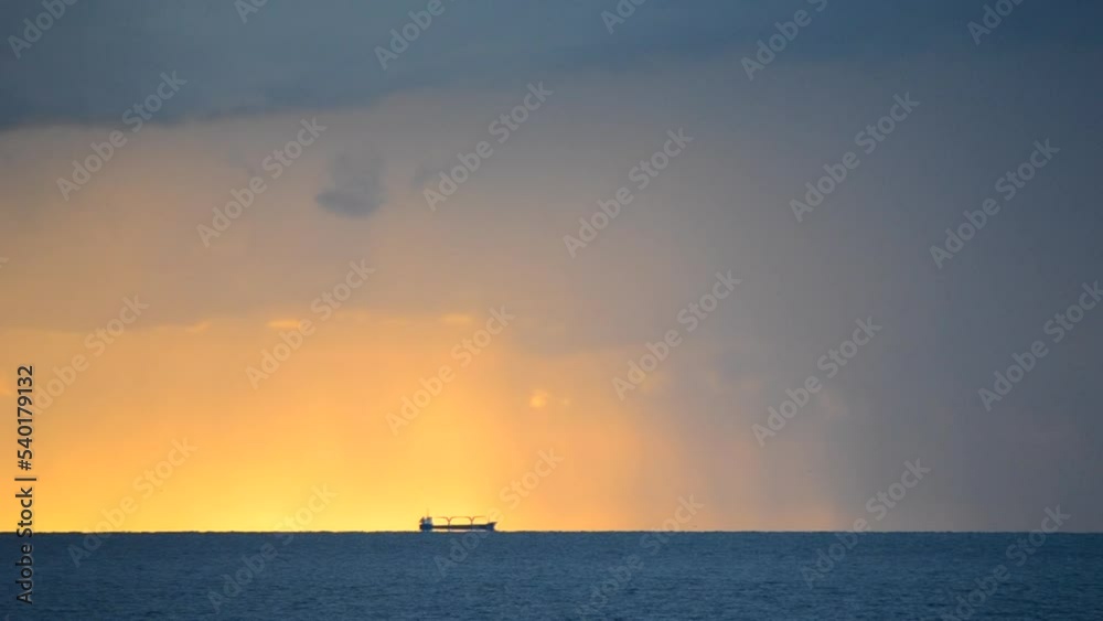 A grain-carrier tanker is far away on the horizon line in the sea against the background of a dawn sunset with falling rain and large dark thunderclouds. Seascape, sea landscape. Natural background A grain-carrier tanker is far away on the horizon line in the sea against the background of a dawn sunset with falling rain and large dark thunderclouds. Seascape, sea landscape. Natural background
