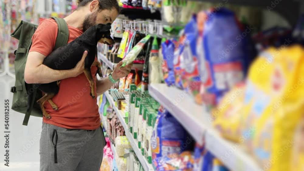 Young man with little lap dog on hands is shopping in pet department of supermarket, 4K footage.