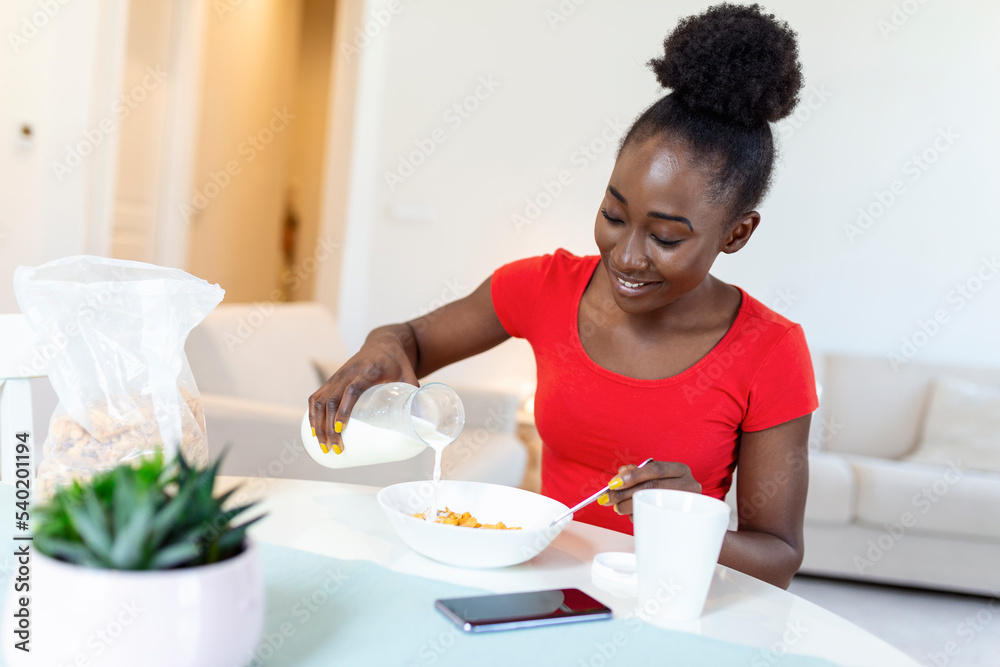 Smiling young African American woman pours corn flakes in plate with milk. The girl has a healthy breakfast on stylish cozy home at the morning while checking her email on laptop.