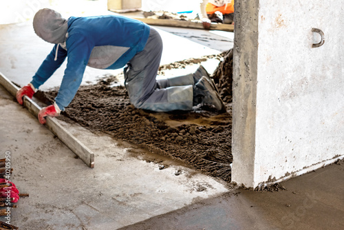 Close up of man builder placing screed rail on the floor covered with sand-cement mix at construction site. Male worker leveling surface with straight edge while screeding floor. Blurred background.