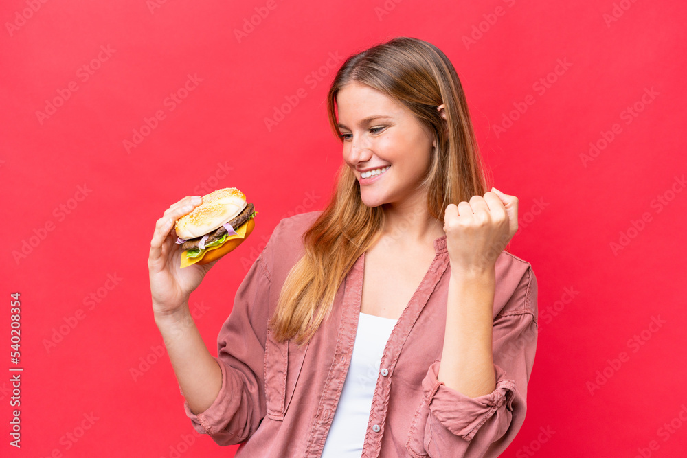 Young caucasian woman holding a burger isolated on red background celebrating a victory Young caucasian woman holding a burger isolated on red background celebrating a victory