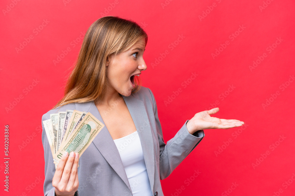 Young business caucasian woman holding money isolated on red background with surprise expression while looking side Young business caucasian woman holding money isolated on red background with surprise expression while looking side