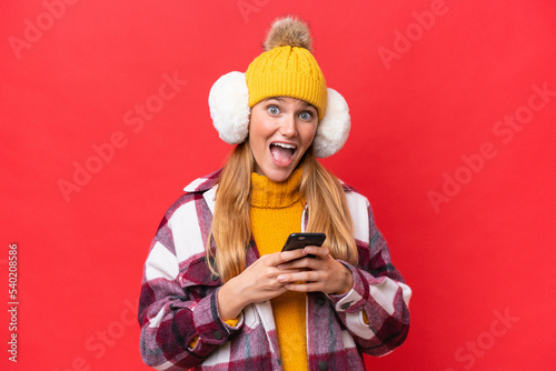 Young beautiful woman wearing winter muffs isolated on red background surprised and sending a message