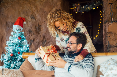 Christmas gifts exchange tradition concept. Happy couple enjoy xmas time holiday at home sharing presents. Woman doing a surprise to her husband man giving him a christmas gift from back. Joy people photo
