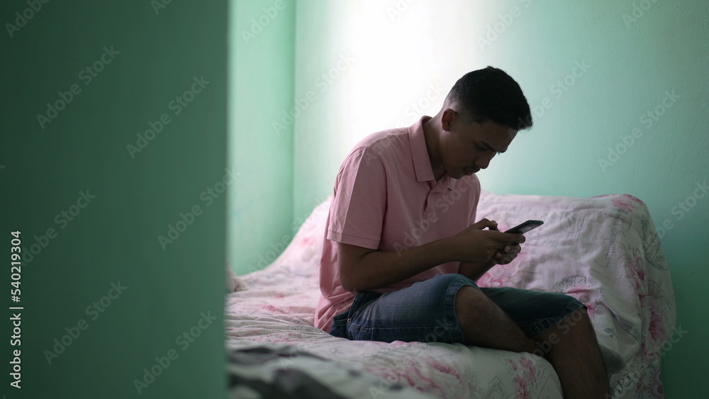 One young hispanic man looking at cellphone device sitting on bed inside bedroom One young hispanic man looking at cellphone device sitting on bed inside bedroom