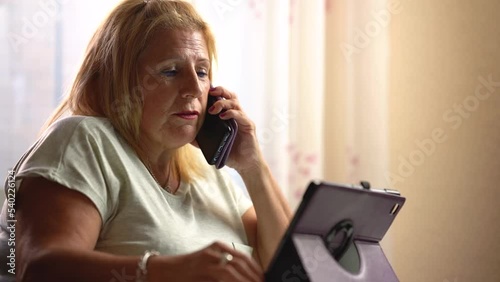 Older woman talking with her smartphone and using the tablet at home - concept of technology and older people