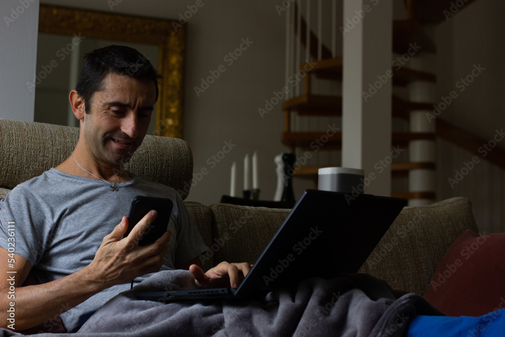 Smiling man using laptop and cellphone sitting on couch covered with blanket. Domestic expenses control, work from home, low key concepts Smiling man using laptop and cellphone sitting on couch covered with blanket. Domestic expenses control, work from home, low key concepts