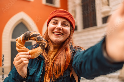 Fall tourist woman in a bright hat and autumn jacket holding baked obwarzanek traditional polish cuisine snack bagel on old city Market square in Krakow  photo