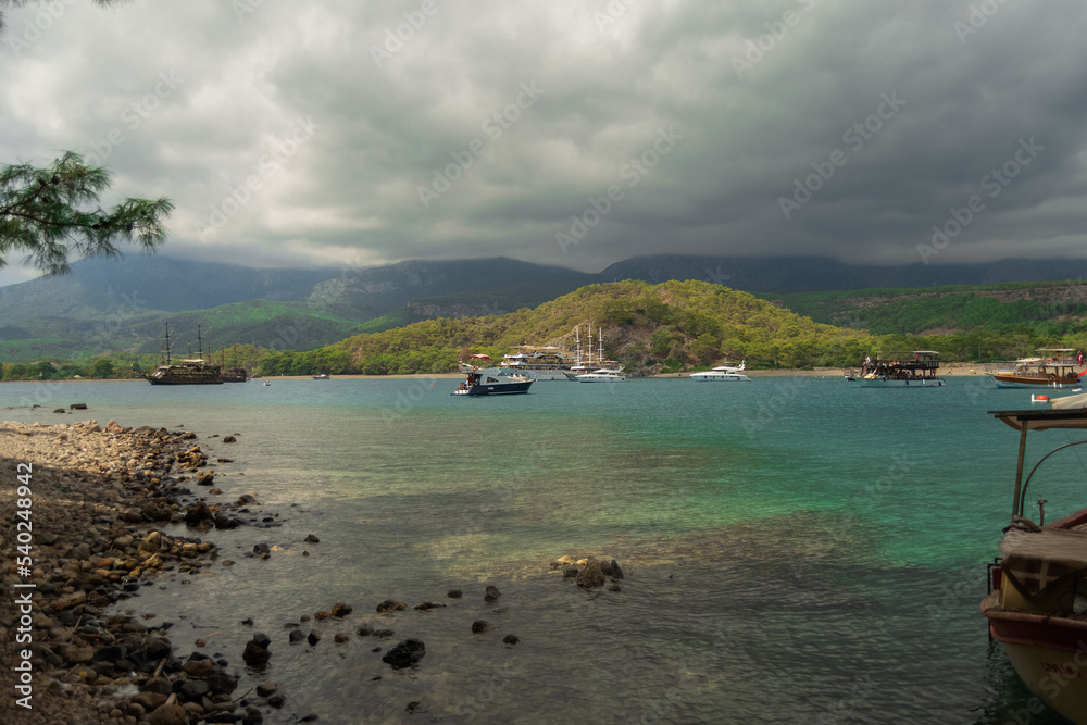 A bay with ships, mountains, clouds, clear blue water and rocks