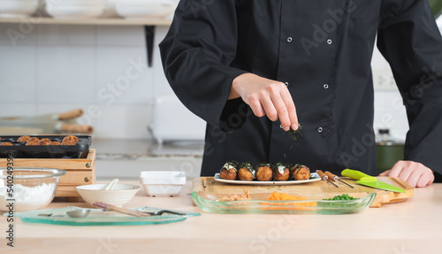 Selective focus on hand of anonymous young chef man, in black uniform, garnishing seaweed on japanese food called takoyaki in plate from hot pan on table at kitchen restaurant