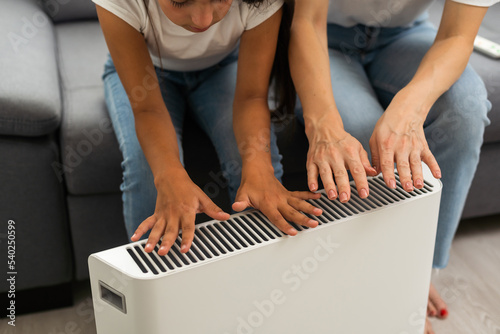 Mother and child warming hands near electric heater at home photo