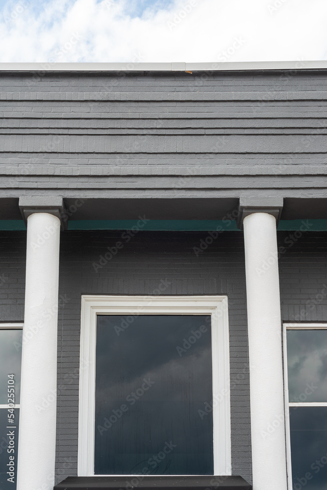 The roof section of an old grey colored cement business building wall. There are two white columns on either side of the door with two glass windows on the outside of the pillars. The sky is grey. The roof section of an old grey colored cement business building wall. There are two white columns on either side of the door with two glass windows on the outside of the pillars. The sky is grey.
