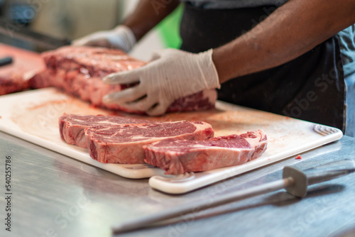 A close up of a large prime rib roast which is sitting on a brown plastic cutting board. The meat has some marbling and multiple ribs. The chef is using a long knife to cut steaks from the fresh meat.