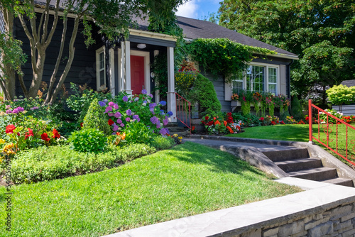 The front yard of a dark blue house with a vibrant red door. The building has a glass window with white trim. The colorful garden has green grass, red, pink, and yellow flowers, shrubs, and trees. 