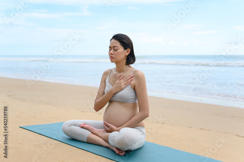 Pregnant woman practices yoga meditation in lotus pose and sits on the peaceful beach with nature scene- Healthy lifestyle concept