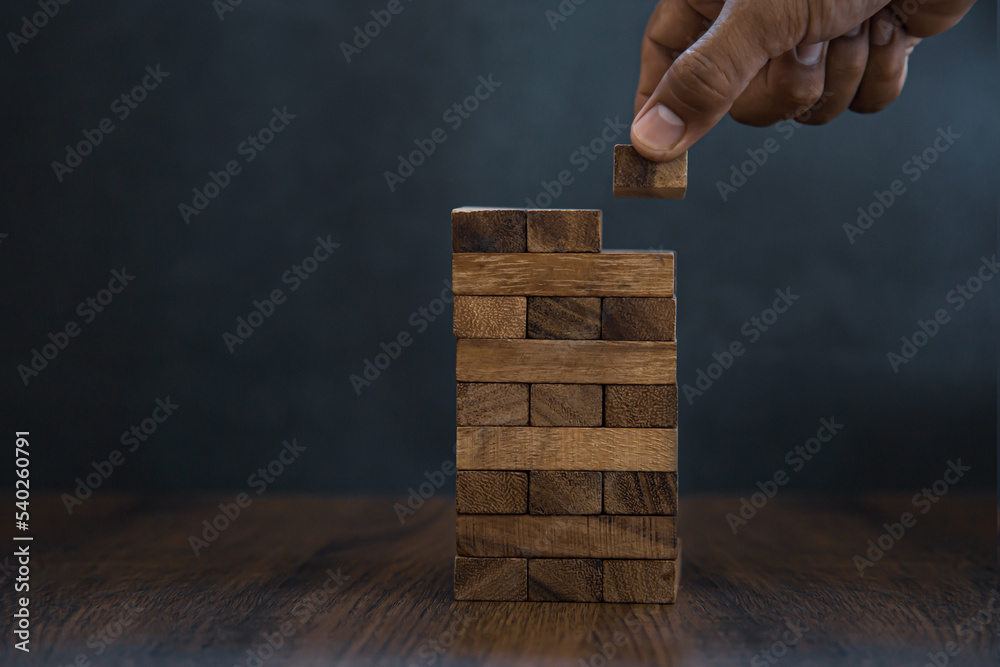 Hand placing wooden block tower stack in pyramid stair step with caution to prevent collapse or crash concepts of financial risk management and strategic planning and business challenge plan. Hand placing wooden block tower stack in pyramid stair step with caution to prevent collapse or crash concepts of financial risk management and strategic planning and business challenge plan.