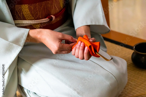 Japanese woman, tea master, Sen Rikyu, hands using an orange purifying cloth to clean a chashaku bamboo matcha tea spoon in Japanese traditional tea ceremony