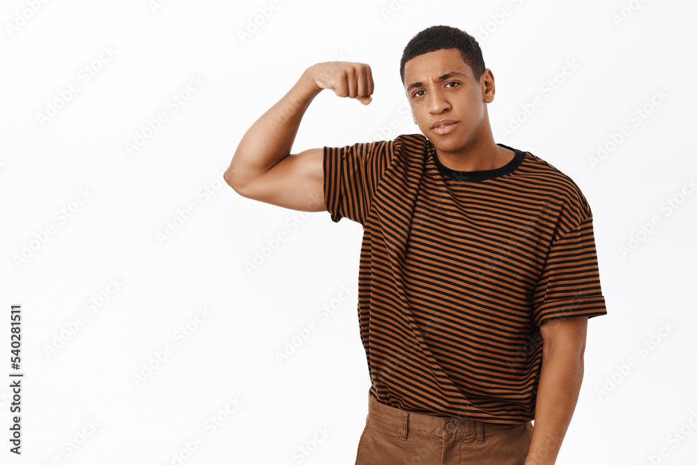 Sport and gym concept. Strong African american man flex biceps, shows muscles, looking confident, standing over white background Sport and gym concept. Strong African american man flex biceps, shows muscles, looking confident, standing over white background