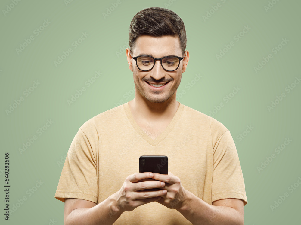 Young handsome man in beige t-shirt and glasses, holding smartphone, looking to screen with smile Young handsome man in beige t-shirt and glasses, holding smartphone, looking to screen with smile