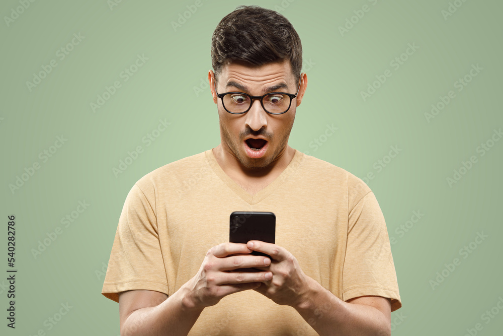 Portrait of young handsome man in t-shirt and glasses, looking at phone screen with shocked face