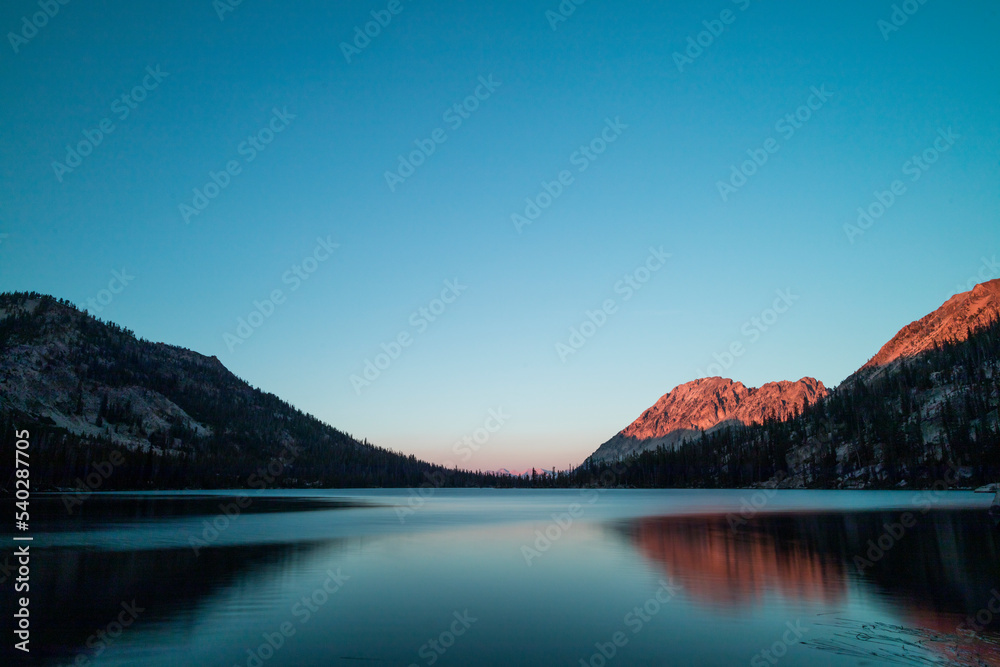 Toxaway Lake, located in Idaho’s Sawtooth Wilderness seen on a summer evening at sunset, with a mountain ridge illuminated reddish orange with the last rays of sunlight. The water reflects the scene.