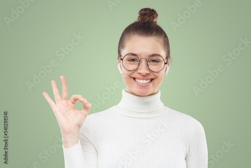 Young woman in glasses showing okay sign to the quality of product, isolated on green background