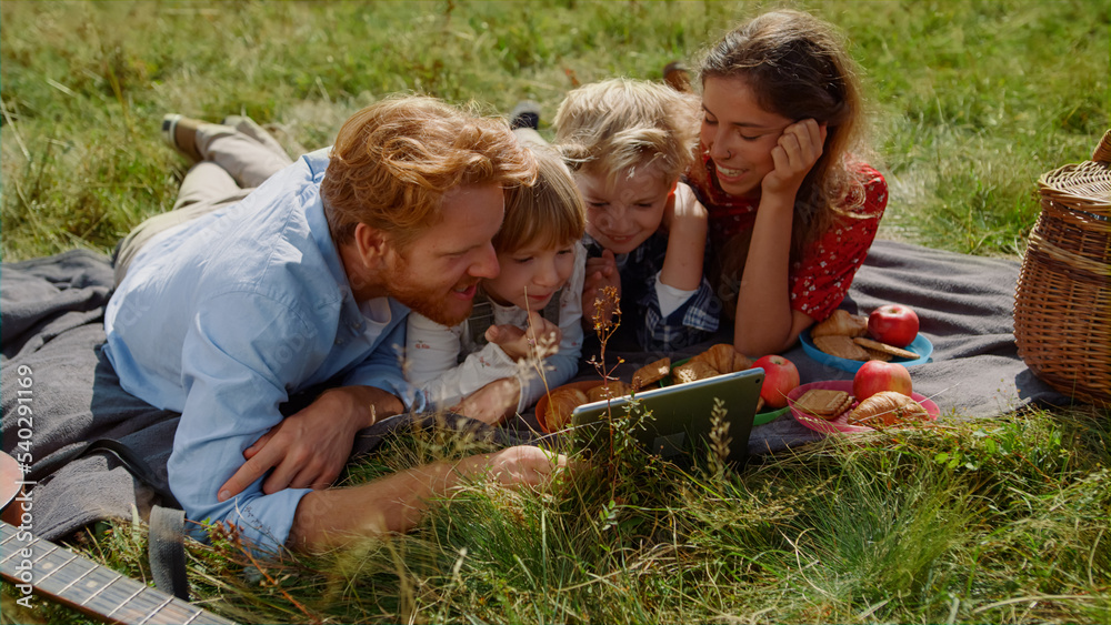 Fototapeta premium Family watching tablet computer on picnic lying green grass. Summer vacation.