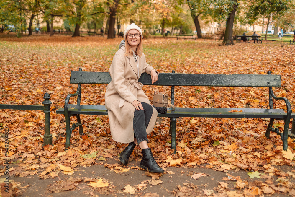 Fall woman in a stylish beautiful warm coat from a knitted hat relaxes on a bench on a warm autumn day Fall woman in a stylish beautiful warm coat from a knitted hat relaxes on a bench on a warm autumn day