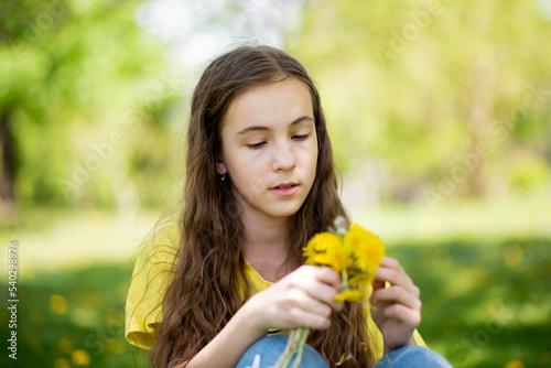 A cute girl on a walk. It s spring outside and the sun is bright. A girl holds dandelions in her hands. She has loose long wavy brown hair. She is dressed in a yellow t-shirt. Childhood. Beauty.