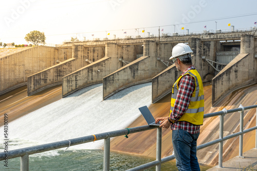 Aืn Engineer standing by the dam. He is wearing a white hard hat and yellow transparent vest.