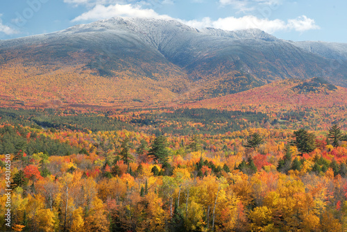Scenic view from Sugar Hill, New Hampshire of treelined valley and hillside covered with vibrant fall foliage and dusting of snow atop summit of Mount Lafayette in Franconia Notch State Park.