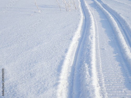 Track of traces from a snowmobile in drifts of white snow. Nature and outdoor on a winter sunny day. Background or backdrop with snowy field