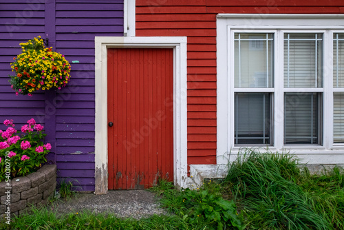 A purple exterior clapboard wooden wall with a hanging flower basket. There s a red door joining the purple wall to a red clapboard wall with a double hung window. There are a flower box and grass too
