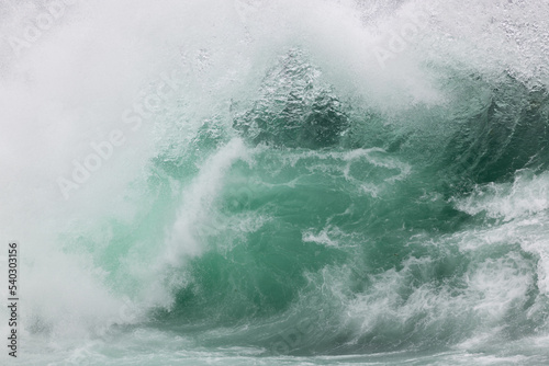 An angry teal green color massive rip curl of a wave as its barrel rolls along the ocean. The white mist and foam from the wave are foamy and fluffy. The ocean spray is coming off the top of the wave