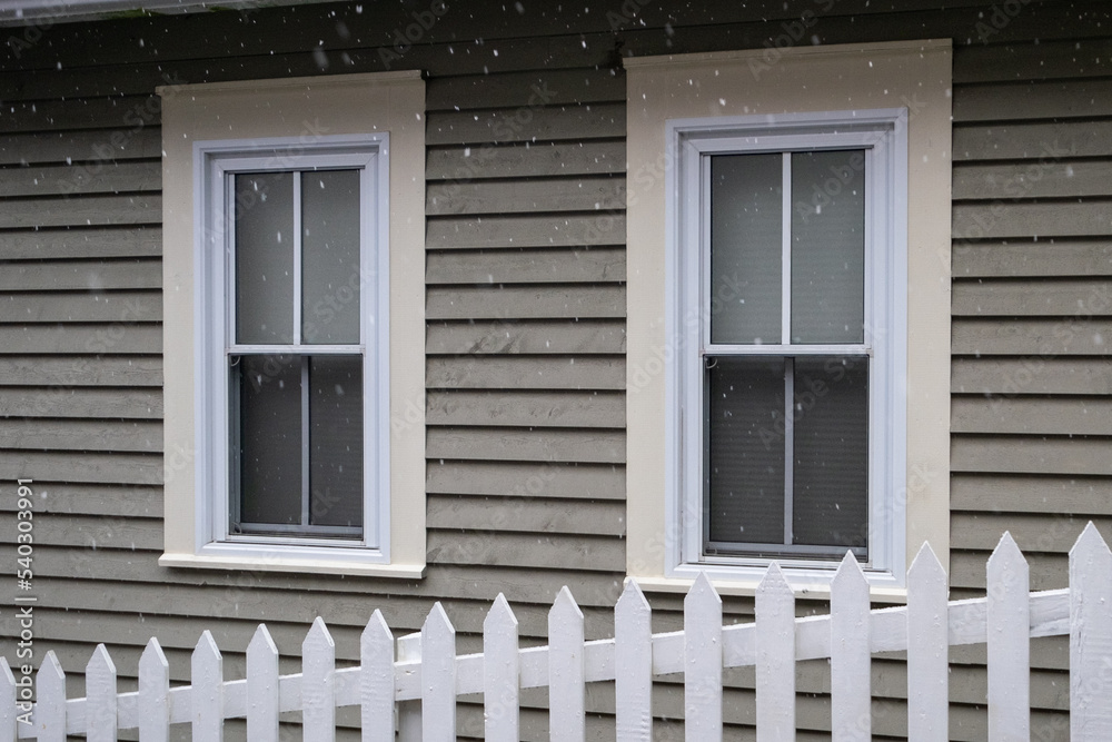Fototapeta premium White picket fence on a diagonal in the foreground with a tan colored house in the background. There are two white double hung windows with beige trim on the exterior wall of the country style home. 