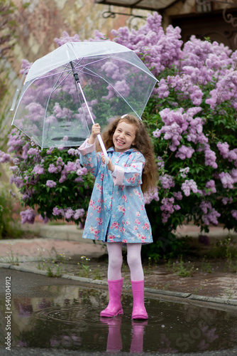 A sweet girl of five years with a transparent umbrella against the background of blooming lilacs enjoys spring and has a fun. Pastel colors. Spring. Childhood. Beauty.