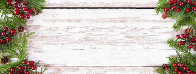 Winter double border with evergreen branches, red berries and pine cones. Overhead view on a rustic white wood banner background with copy space.