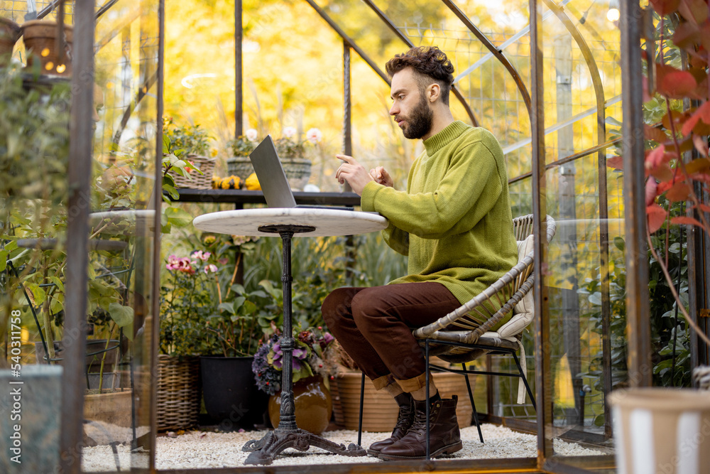 Man works on laptop while sitting by the round table in glasshouse with plants and flowers at backyard. Work from home at cozy atmosphere on nature