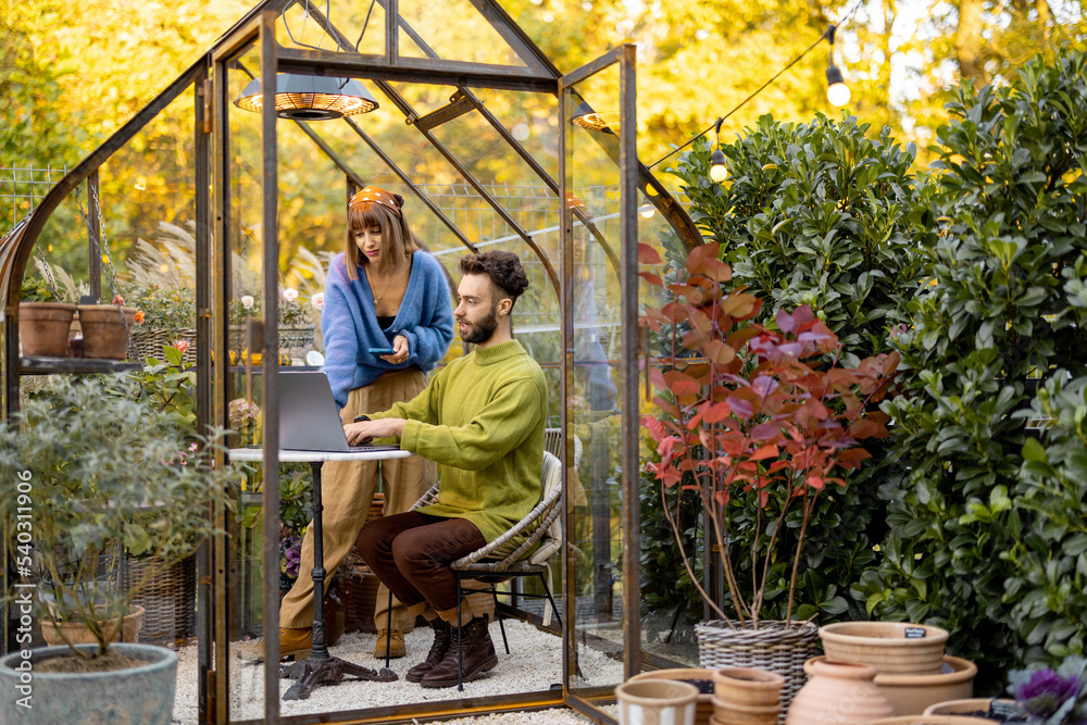 Man and woman have a conversation while work on laptop by the round table in glass house with plants and flowers at backyard. Work from home at cozy atmosphere on nature Man and woman have a conversation while work on laptop by the round table in glass house with plants and flowers at backyard. Work from home at cozy atmosphere on nature