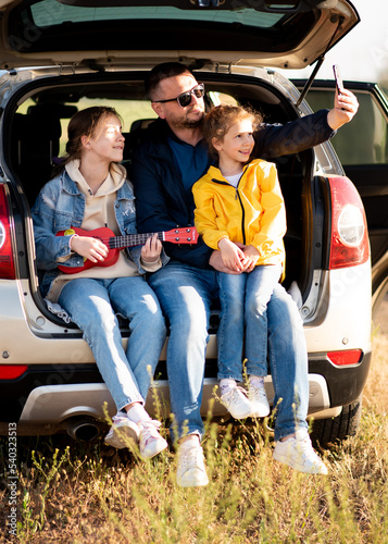 A dad with two daughters in the car. They sit in the open trunk of the car. The girl plays the ukulele. Dad takes a selfie. Communication. Family vacation.