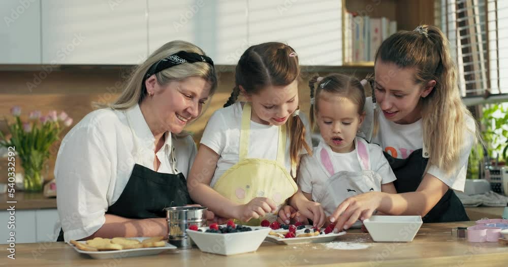 Happy mother's day. delighted middle aged grandmother with daughter and adorable preschooler granddaughters dressing homemade cookies with berries standing at table in modern light kitchen in aprons. Happy mother's day. delighted middle aged grandmother with daughter and adorable preschooler granddaughters dressing homemade cookies with berries standing at table in modern light kitchen in aprons.