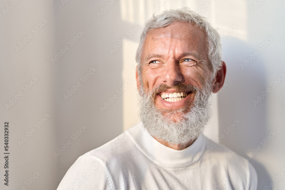 Obraz premium Smiling cheerful bearded mature older fashion gray-haired business man, happy old senior businessman looking away at window laughing standing at white wall lit with sunlight rays. Headshot