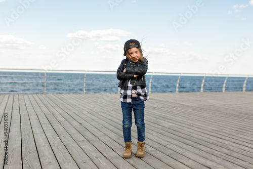 Full-lenght photo of stylish little kid in jeans, leather jacket and cap crossed arms on waist and standing on wooden pier against sea in warm spring day.