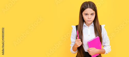 Serious frown child hold school book and pen yellow background. Portrait of schoolgirl student  studio banner header. School child face  copyspace.