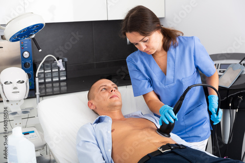 Young male patient getting fat reductive skin lifting body treatment by female cosmetologist in clinic photo